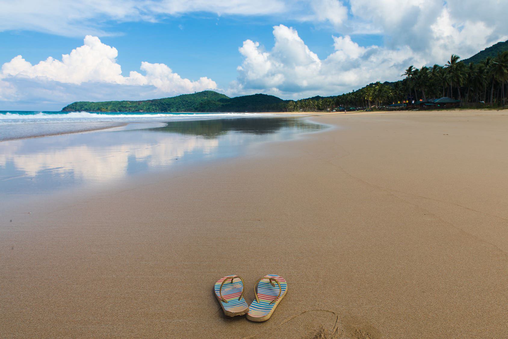 pair of flipflops at Nacpan Beach with cloudy sky and tropical trees on background in El Nido, Palawan province, Philippines